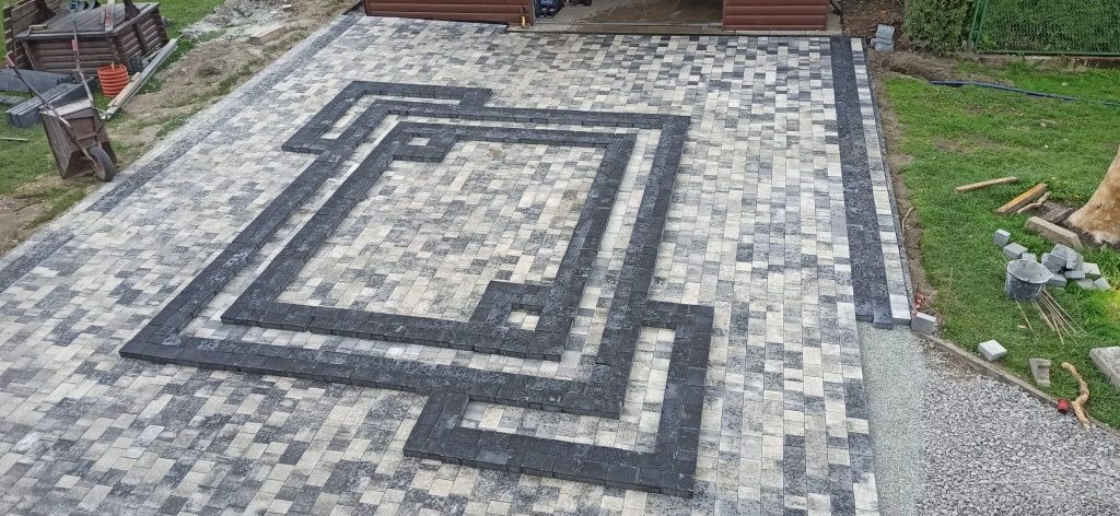 Ułożona kostka brukowa z dekoracyjnym wzorem geometrycznym z ciemnych kostek, widok z góry, teren wokół częściowo zagospodarowany, w tle widoczny garaż.