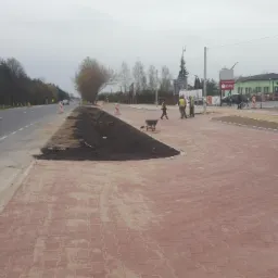 Nowa czerwona kostka brukowa ułożona przy drodze, z przygotowanym terenem pod nasadzenia i pracownikami w tle, widoczny pomarańczowy samochód serwisowy.