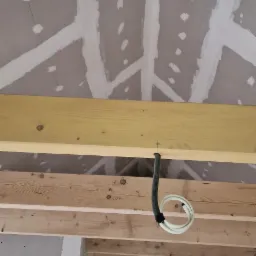 Sufit z płyt kartonowo-gipsowych w trakcie montażu, widoczne drewniane belki konstrukcyjne, szpachlowane łączenia płyt i przew&oacute;d elektryczny zwisający z sufitu, drabina w tle.