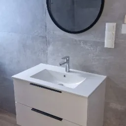 Nowoczesna łazienka z umywalką nablatową na szafce z dwiema szufladami z czarnymi uchwytami, okrągłe lustro zawieszone na sznurze, szare płytki na ścianach i drewnopodobna podłoga.