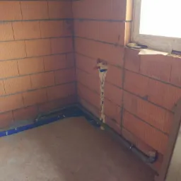 Wewnętrzne ściany z cegły dziurawki z częściowo poprowadzonymi rurami instalacji wodnej i kanalizacyjnej, zabezpieczone pianką montażową przy ścianie, widoczne elementy okna i podłogi w surowym...