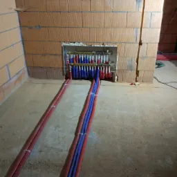 Rozdzielacz ogrzewania podłogowego w trakcie montażu, widoczne rury doprowadzające i odprowadzające wodę, całość umieszczona w wycięciu w ścianie z cegły.