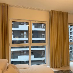 Studio Aranżacji Okien - Beżowe, gładkie zasłony na szynach sufitowych w nowoczesnym salonie z widokiem na budynek mieszkalny. Częściowo widoczny kremowy narożnik.