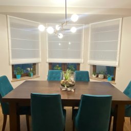 Studio Aranżacji Okien - Jadalnia z drewnianym stołem, turkusowymi krzesłami i trzema oknami z białymi roletami rzymskimi. Na parapecie doniczki z roślinami. Nad stołem nowoczesna lampa.