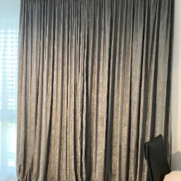 Eleganckie, grafitowe zasłony z aksamitnej tkaniny, upięte w regularne fałdy, wpadają miękko na podłogę. W tle okno z białymi roletami.