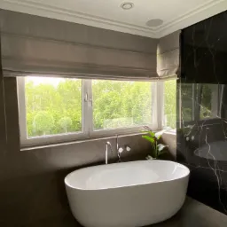 Elegancka łazienka z wolnostojącą wanną przy oknie z szarą roletą rzymską i widokiem na zieleń. Marmurowe ściany i nowoczesna armatura.