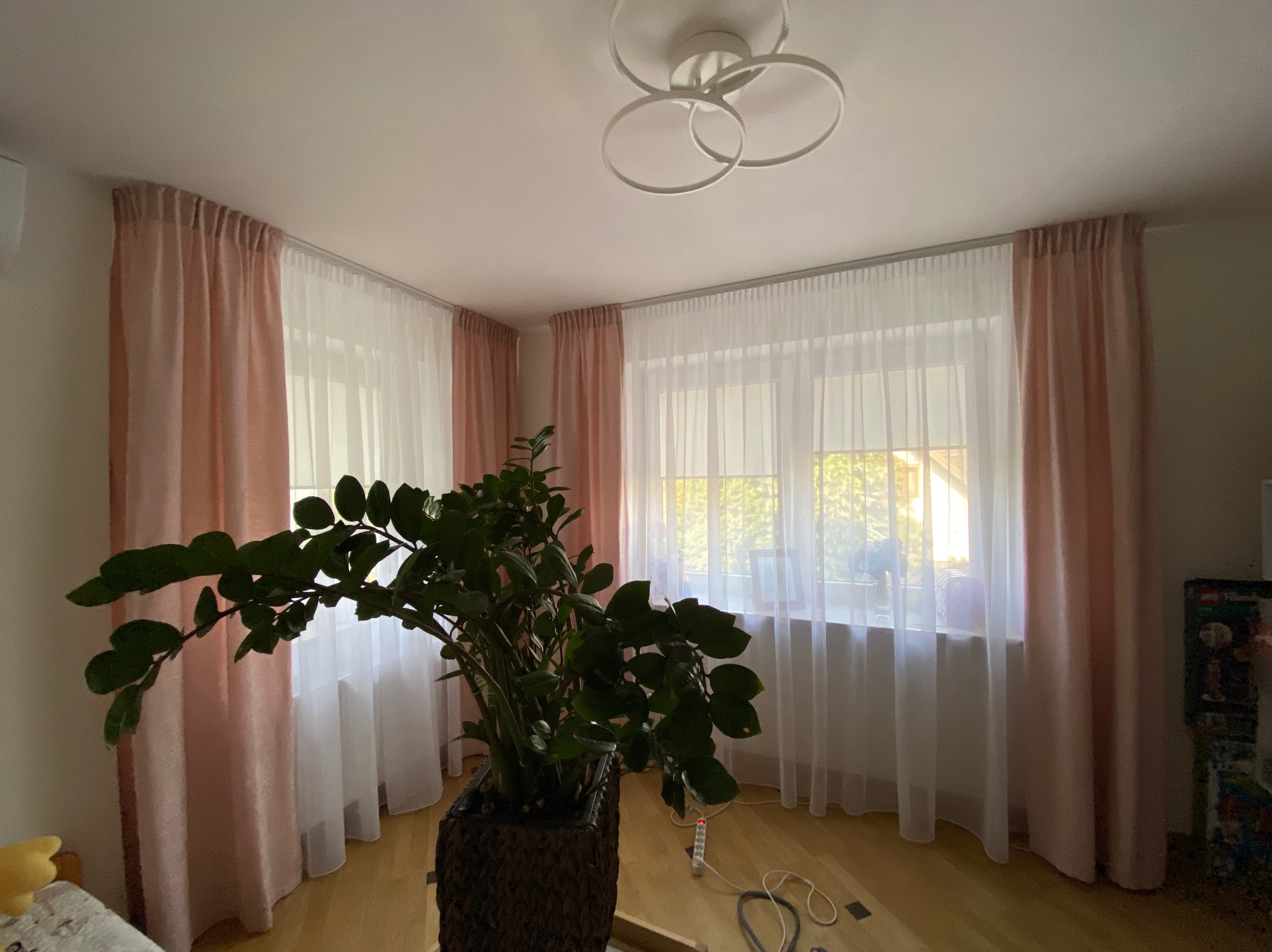 Jasny pokój z oknem przysłoniętym delikatnymi, różowymi zasłonami i białymi firanami. Na pierwszym planie duża roślina doniczkowa.