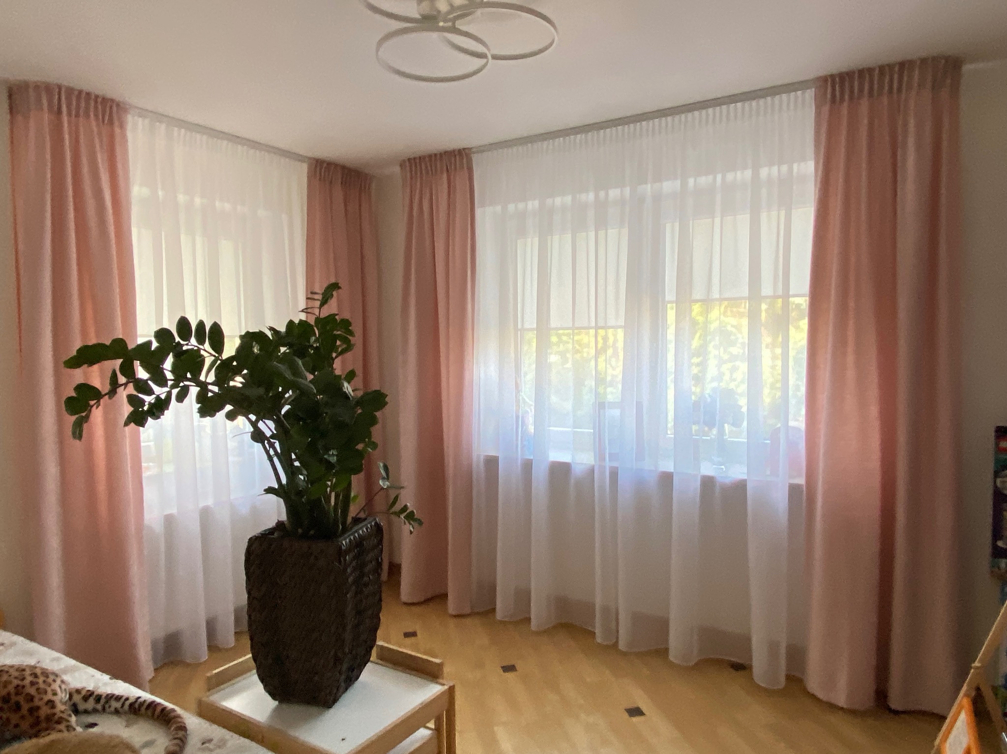 Jasny pokój z oknami przysłoniętymi białymi firankami i pudrowo-różowymi zasłonami. W tle widoczny fragment wnętrza i roślina doniczkowa na stoliku.