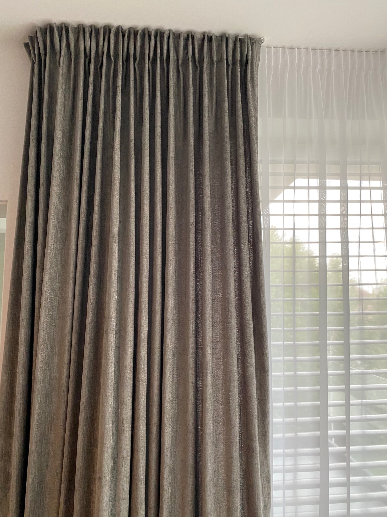 Drapowana, szara zasłona obok białej firanki z żaluzjami w tle, tworząca eleganckie i funkcjonalne okienne wykończenie.