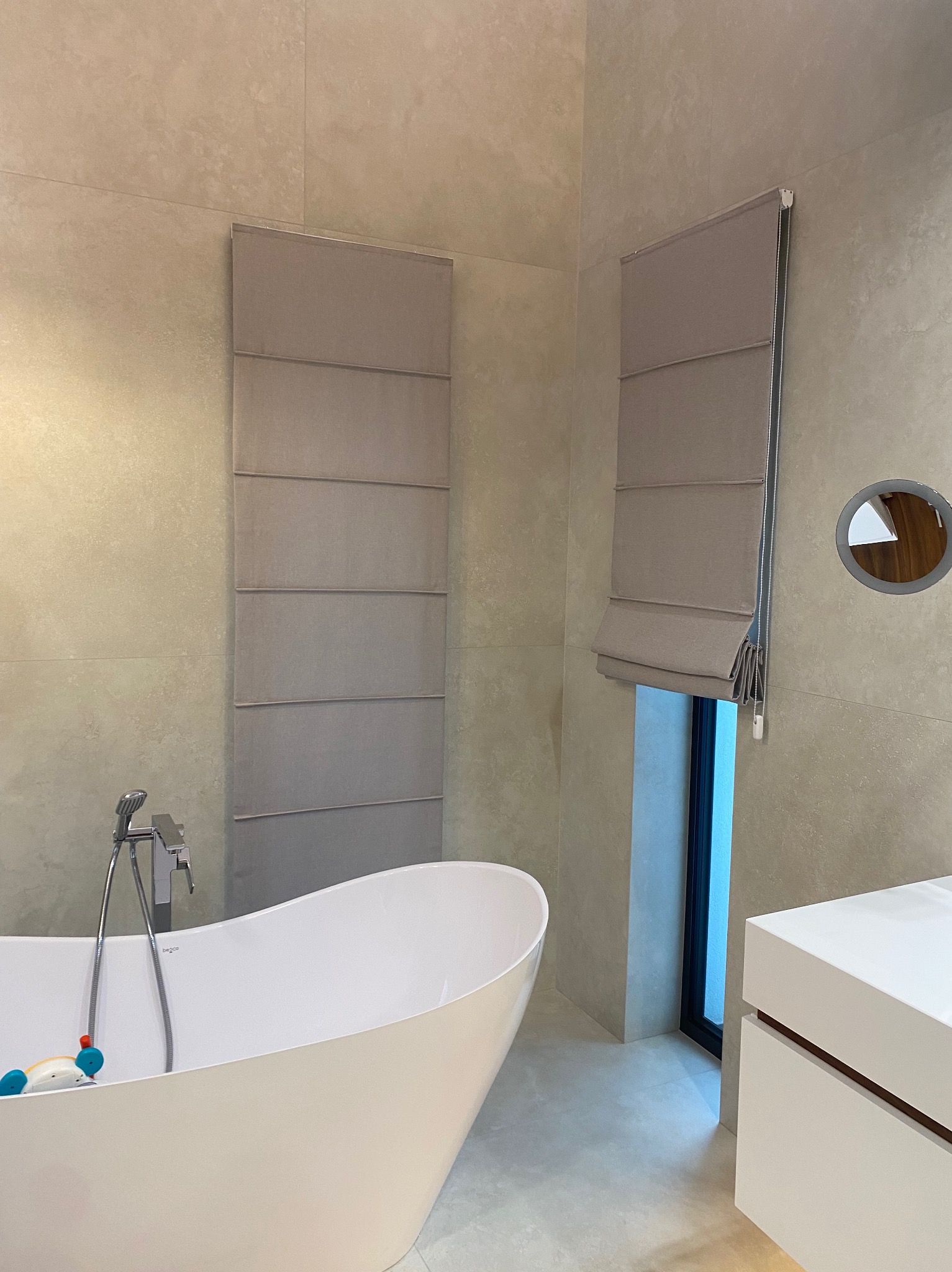 Elegancka łazienka z beżowymi, rzymskimi roletami, białą wanną wolnostojącą i minimalistyczną szafką umywalkową. Okrągłe lustro z podświetleniem.