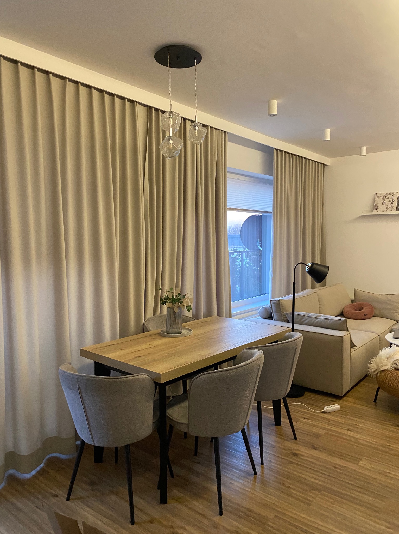 Beżowe, gładkie zasłony w salonie z drewnianym stołem i szarymi krzesłami. Widok na okno i część sofy. Neutralna kolorystyka wnętrza.