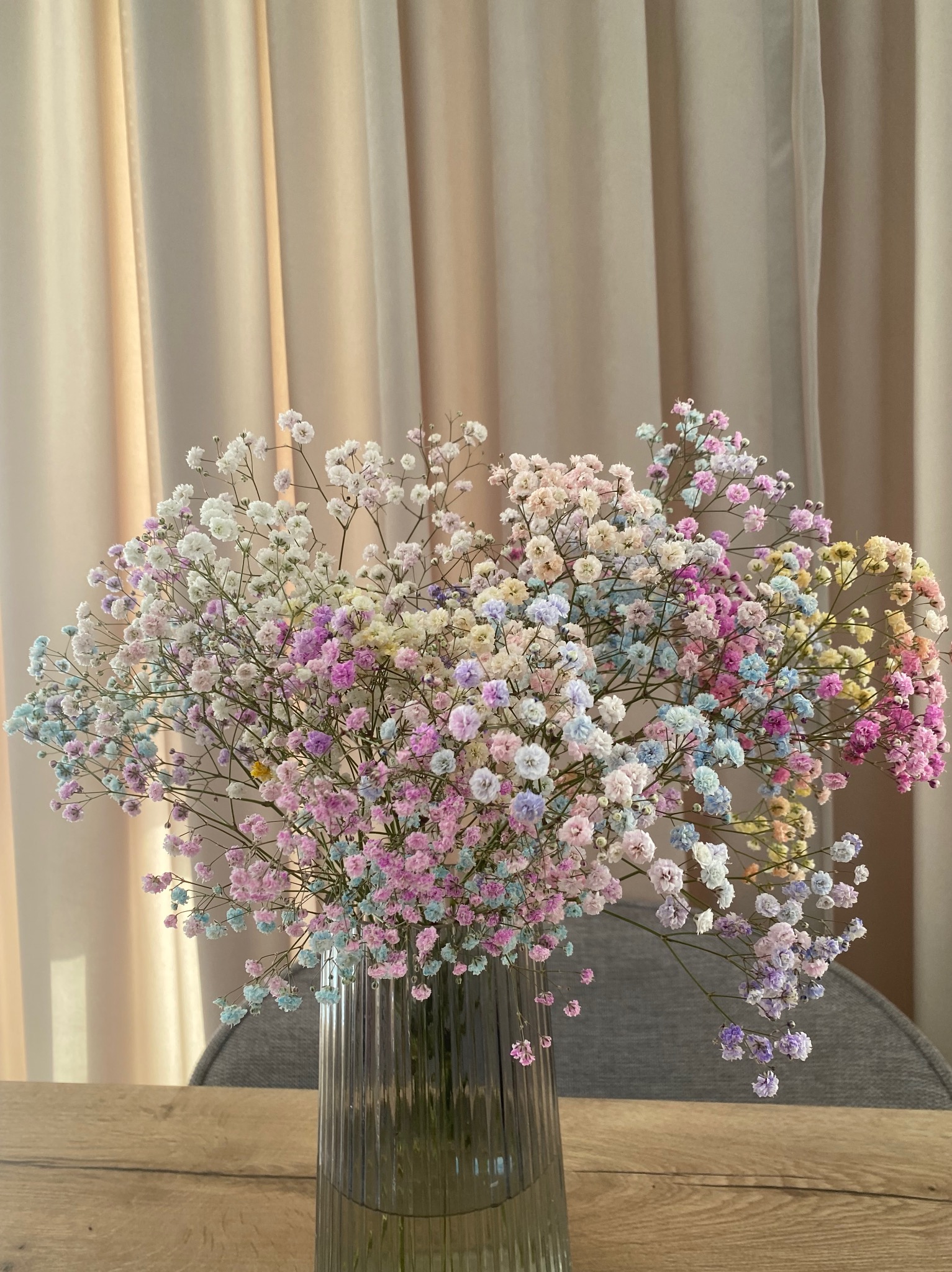 Kolorowy bukiet gipsówki w szklanym wazonie na drewnianym stole, z beżową zasłoną w tle. Pastelowe odcienie kwiatów tworzą delikatny, dekoracyjny akcent.