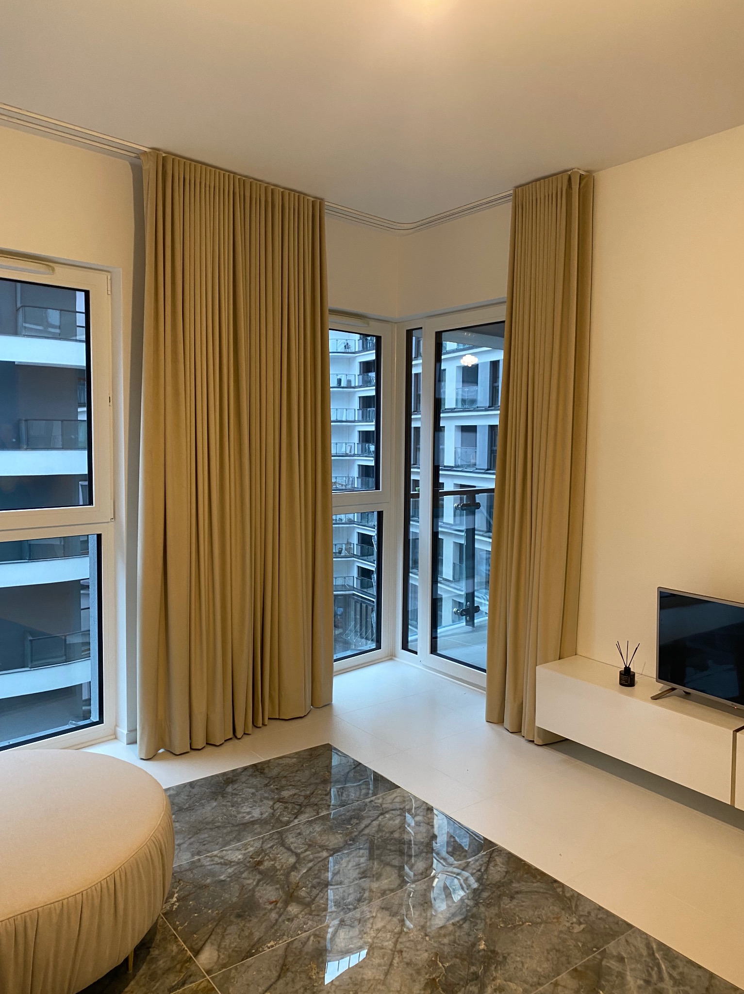 Beżowe, plisowane zasłony w nowoczesnym salonie z dużymi oknami. Widok na apartamentowiec z balkonu. Marmurowa podłoga i minimalistyczna szafka RTV.