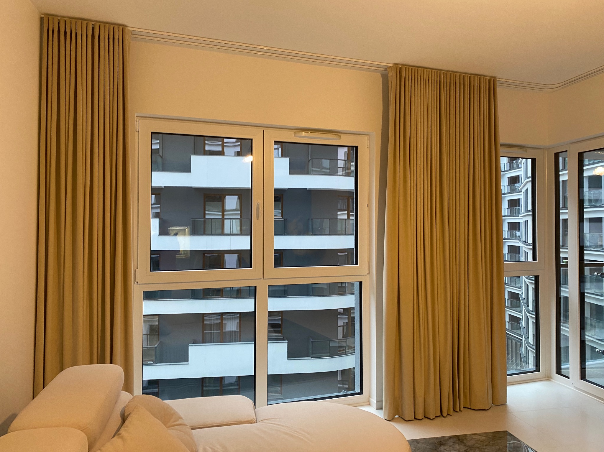 Beżowe, gładkie zasłony na szynach sufitowych w nowoczesnym salonie z widokiem na budynek mieszkalny. Częściowo widoczny kremowy narożnik.
