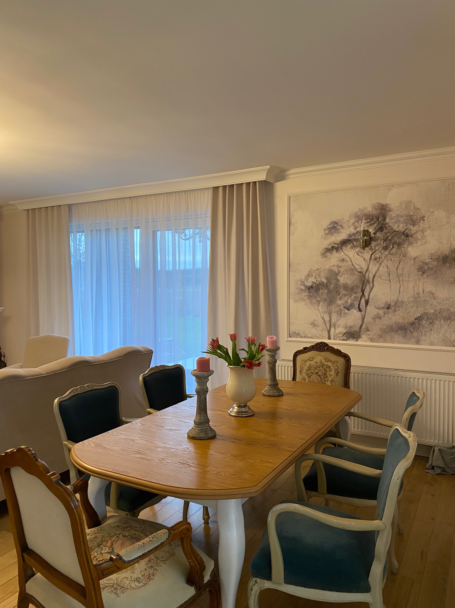 Stylowa aranżacja jadalni z drewnianym stołem, kwiatami w wazonie i dekoracyjnymi świecznikami. W tle zasłony i obraz z motywem drzewa.