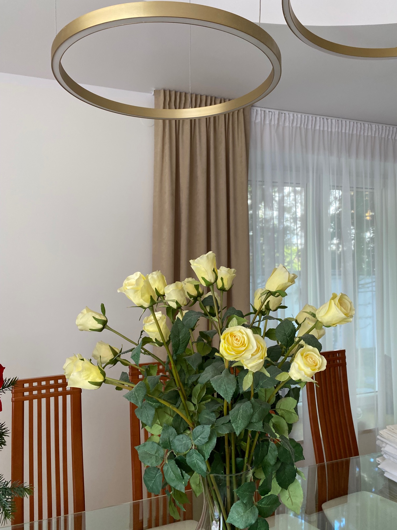 Beżowe zasłony i firany w oknie, nad stołem złote, okrągłe lampy. Na stole wazon z bukietem żółtych róż. Widoczne krzesła i fragment świątecznej dekoracji.