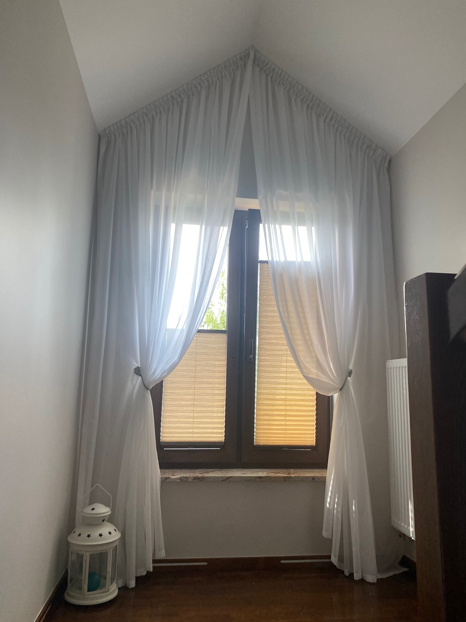 Okno na poddaszu z jasnymi, zwiewnymi zasłonami, upiętymi ozdobnymi klamrami. W tle widoczne rolety plisowane. U dołu dekoracyjna latarenka.