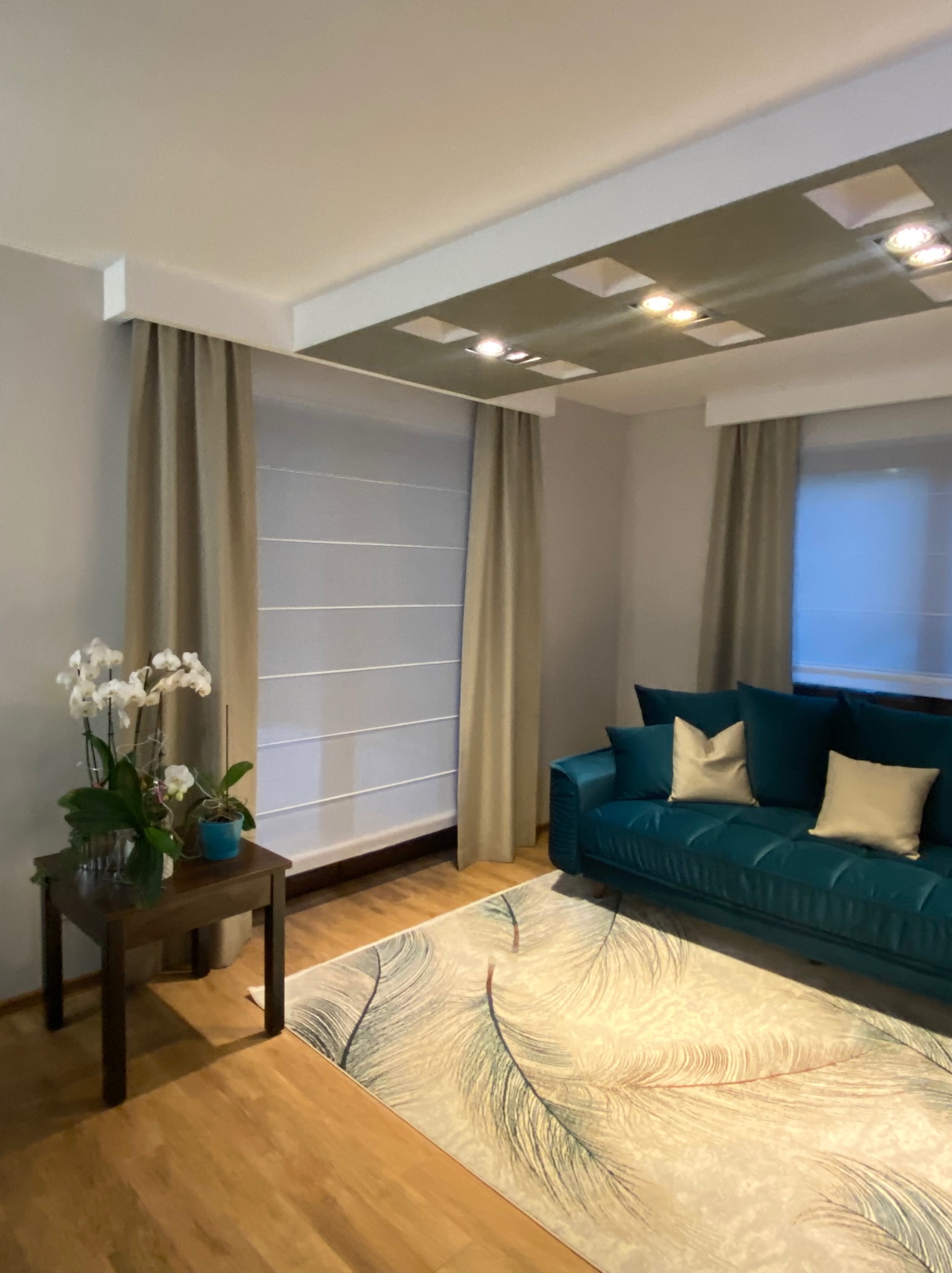 Stylowy salon z turkusową sofą, beżowymi zasłonami i roletą rzymską. Na pierwszym planie drewniany stolik z orchideą. Dywan z motywem piór dodaje wnętrzu charakteru.