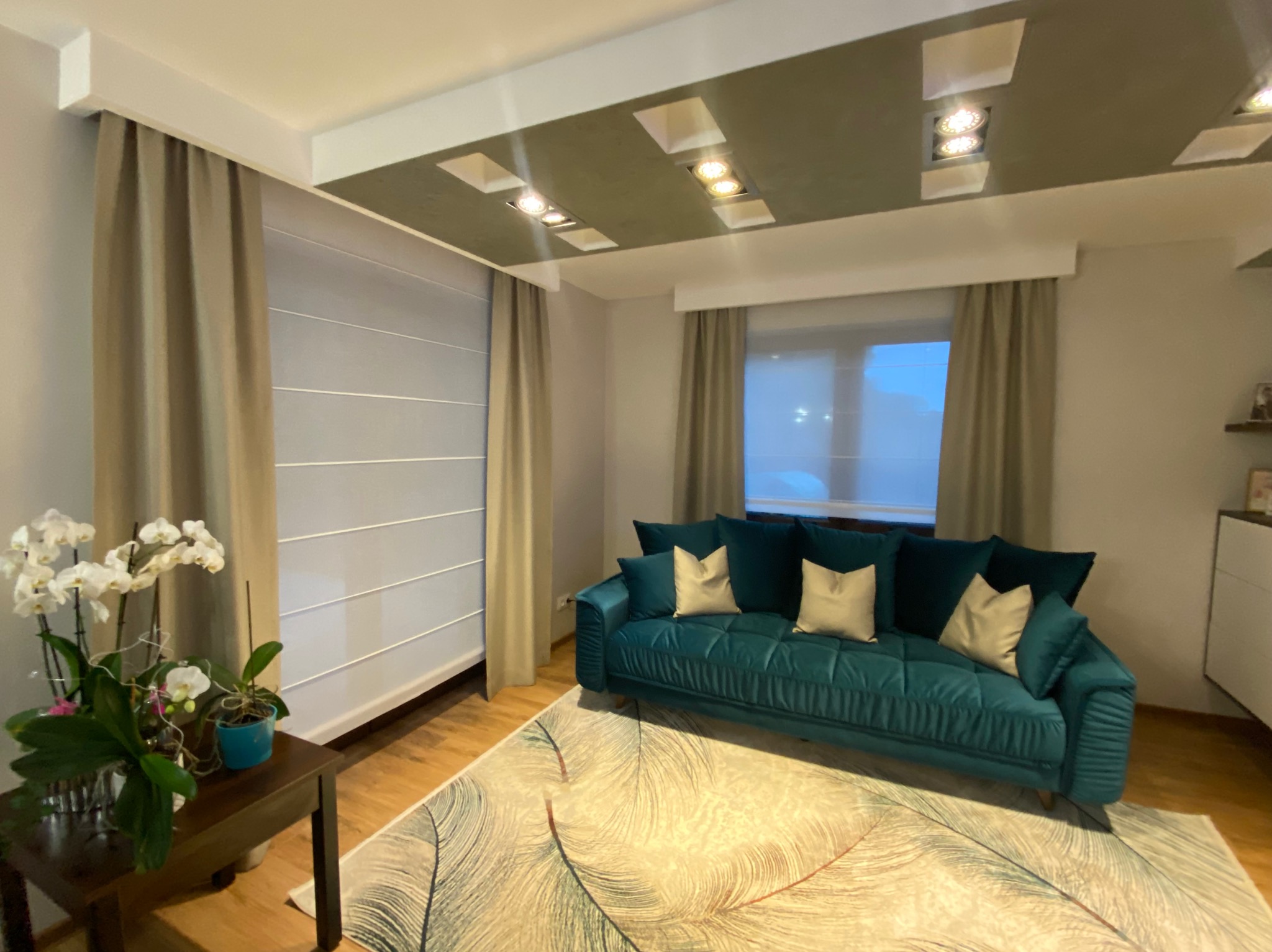 Elegancki salon: turkusowa sofa z poduszkami, wzorzysty dywan, zasłony i rolety poziome. Oświetlenie sufitowe i kwiaty na stoliku dodają przytulności.