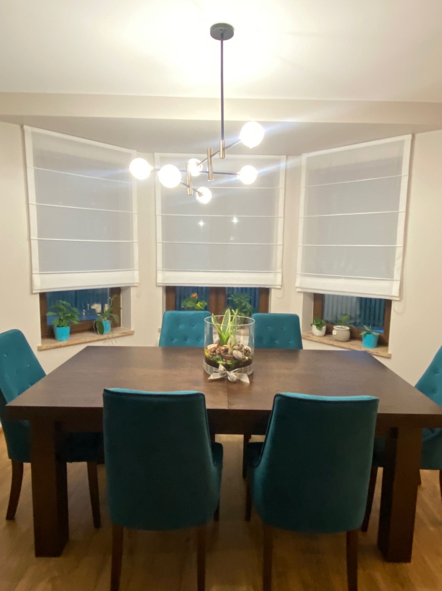 Jadalnia z drewnianym stołem, turkusowymi krzesłami i trzema oknami z białymi roletami rzymskimi. Na parapecie doniczki z roślinami. Nad stołem nowoczesna lampa.