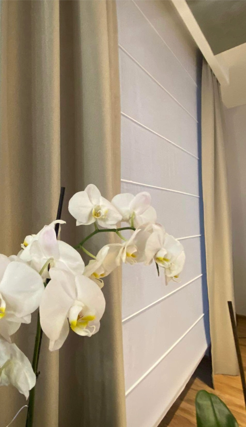 Beżowe zasłony i białe rolety rzymskie z poziomymi listwami, dopełnione kwitnącą orchideą. Subtelna aranżacja okna w neutralnej kolorystyce.