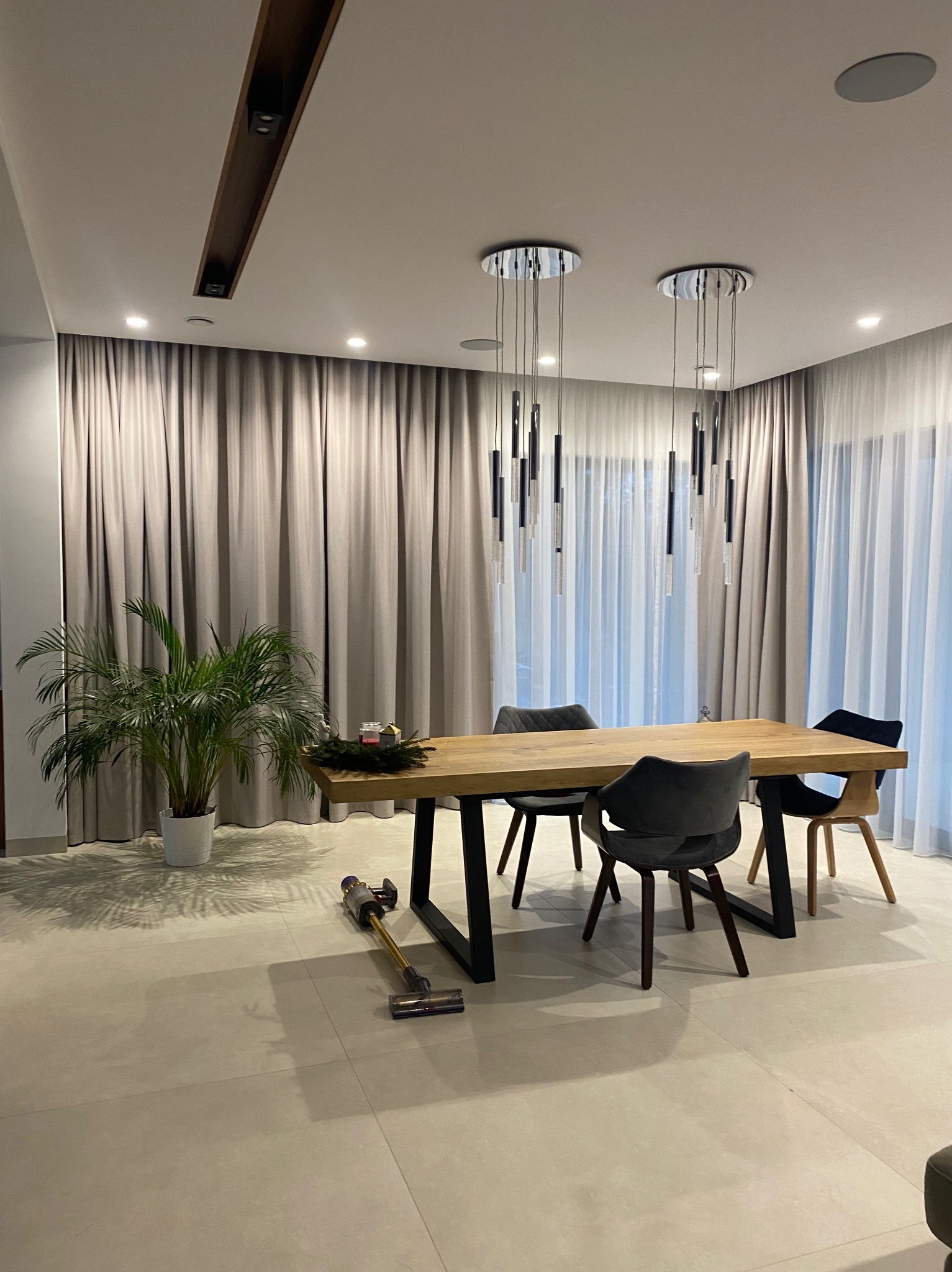 Jadalnia w neutralnej tonacji z drewnianym stołem, szarymi krzesłami i zasłonami. Na podłodze odkurzacz, a w rogu rośliny doniczkowe. Nowoczesne lampy wiszące.