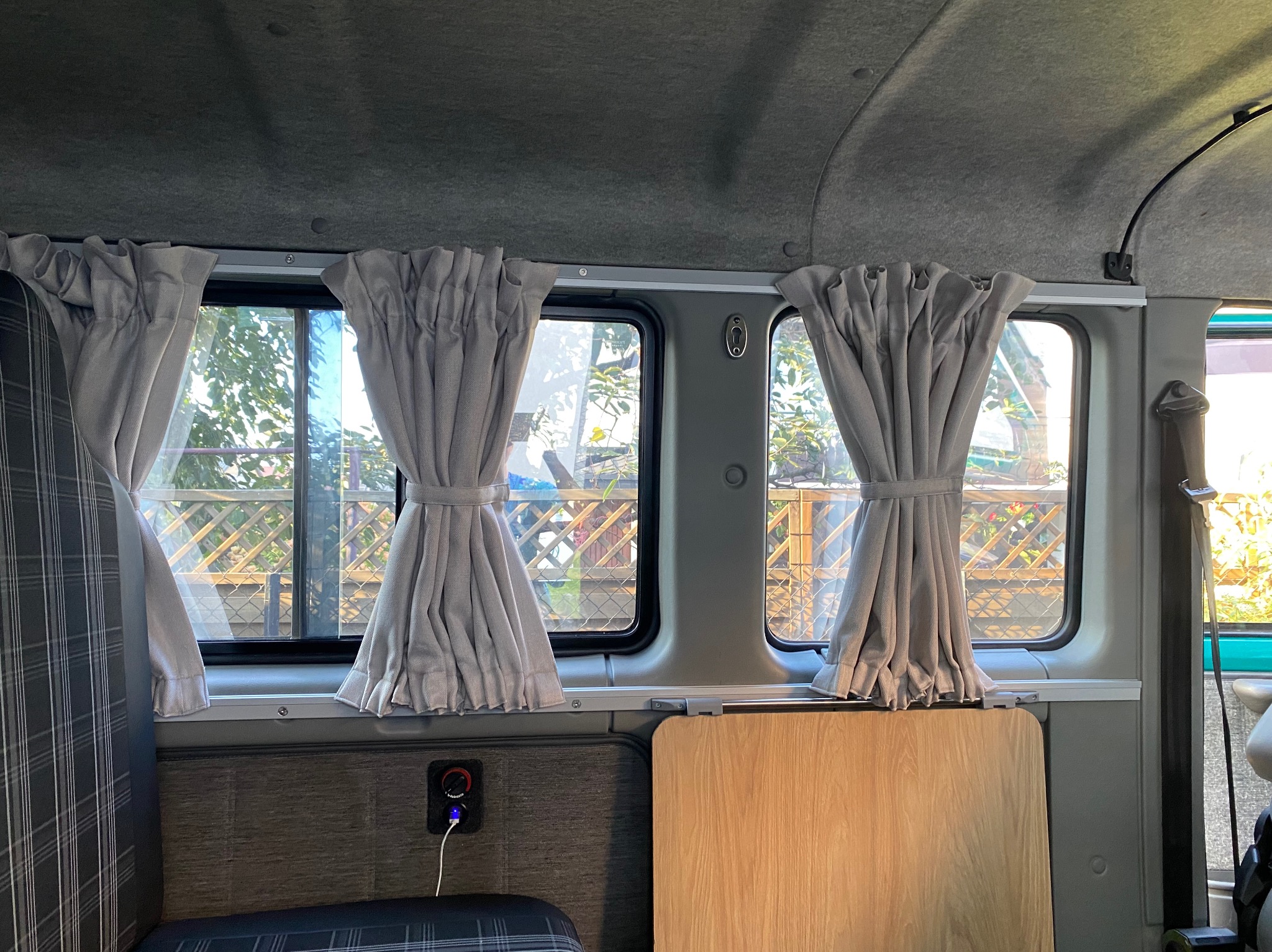 Zasłony w kamperze, upięte w połowie wysokości, w tle widok na zewnątrz przez okna. Wnętrze pojazdu wykończone w szarościach.