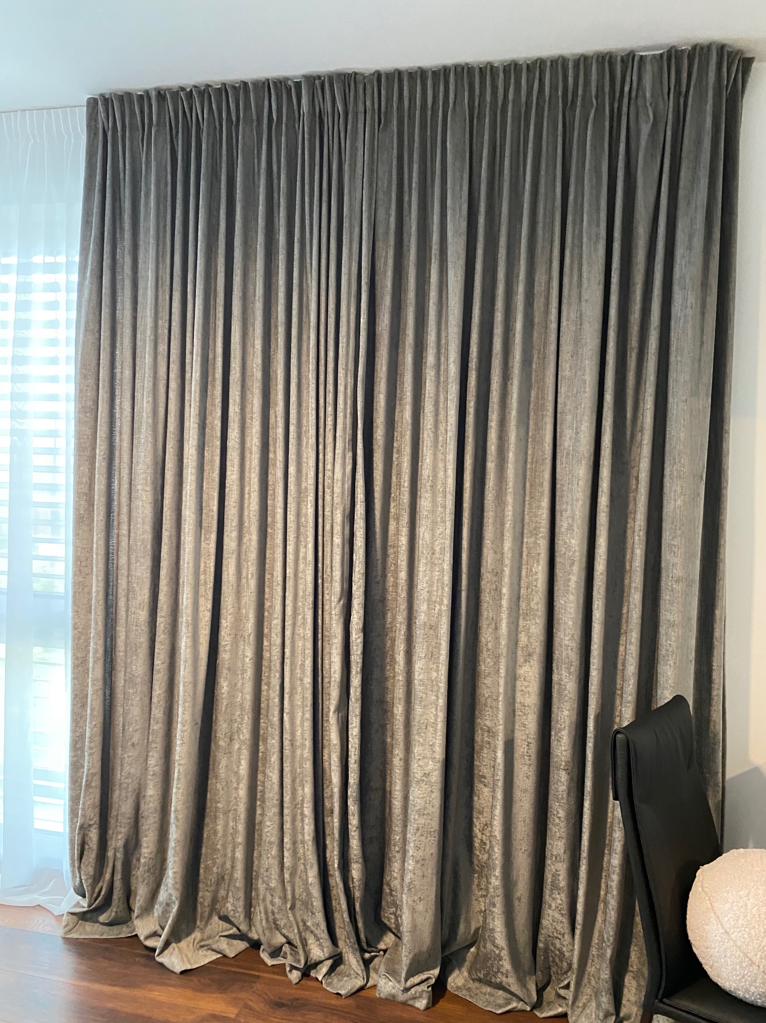 Eleganckie, grafitowe zasłony z aksamitnej tkaniny, upięte w regularne fałdy, wpadają miękko na podłogę. W tle okno z białymi roletami.