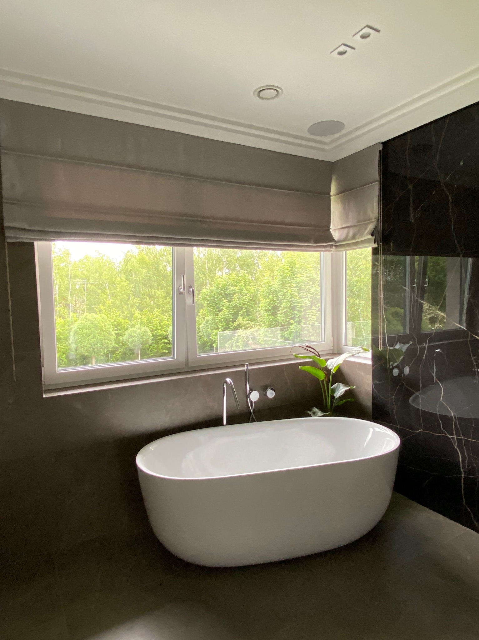 Elegancka łazienka z wolnostojącą wanną przy oknie z szarą roletą rzymską i widokiem na zieleń. Marmurowe ściany i nowoczesna armatura.
