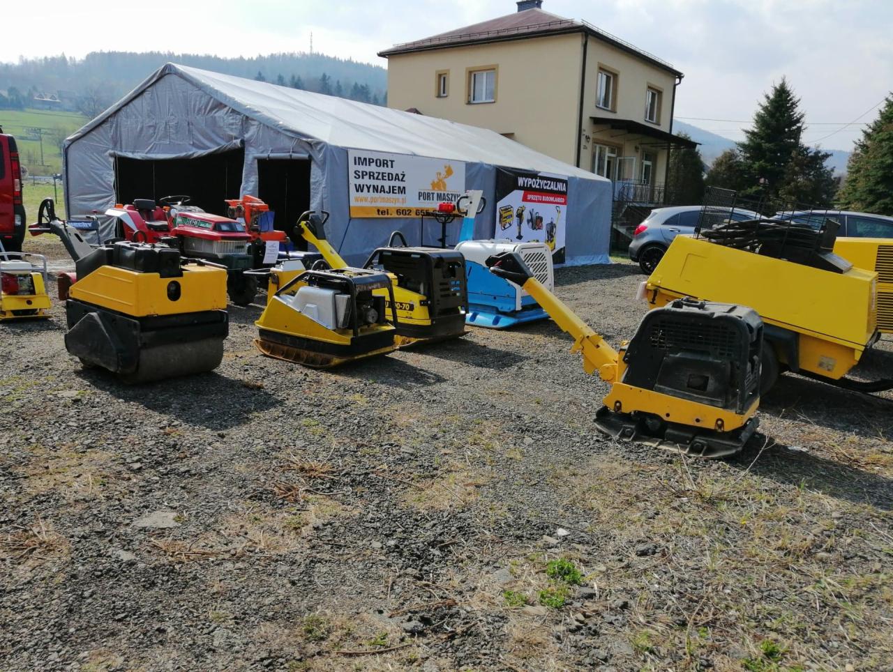 Wystawa żółtych zagęszczarek i walców drogowych na żwirowym placu przed namiotem firmy importującej, sprzedającej i wynajmującej maszyny, w tle budynek i góry.