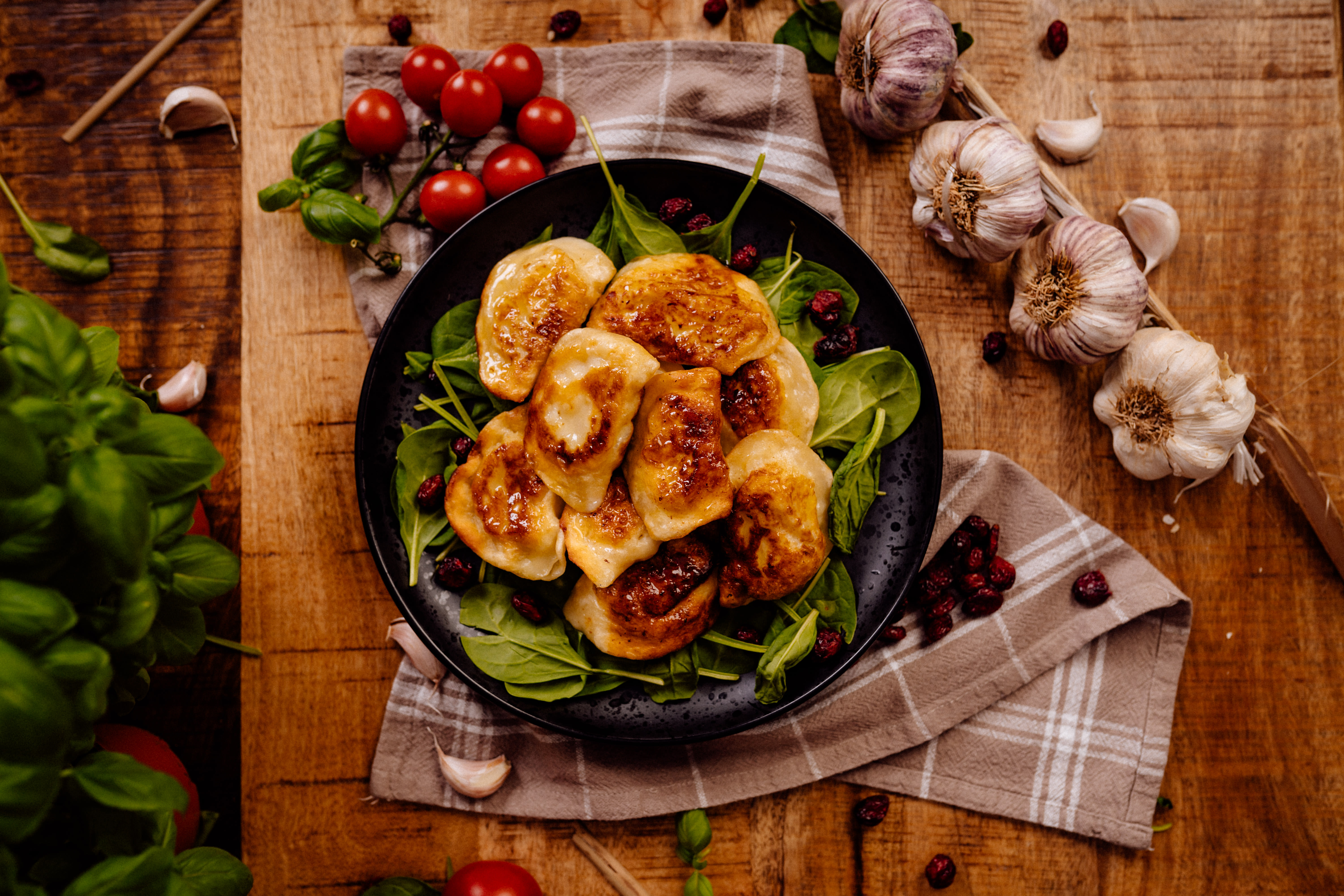Apetycznie wyglądające, smażone pierogi na czarnym talerzu, udekorowane świeżym szpinakiem i żurawiną, otoczone główkami czosnku, gałązkami pomidorków cherry i lnianą ściereczką na rustykalnej...