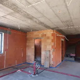 Surowy stan wnętrza z instalacją elektryczną i wodną: białe kable elektryczne na suficie, czerwone rury ogrzewania podłogowego i niebieskie rury wodne na posadzce, ściany z czerwonej cegły dziurawki.