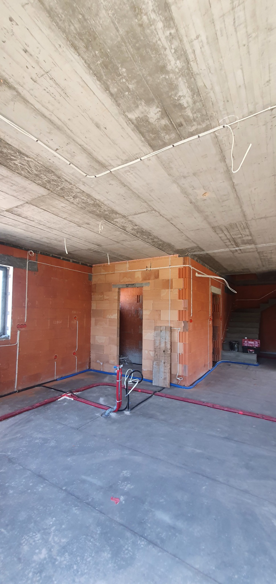 Surowy stan wnętrza z instalacją elektryczną i wodną: białe kable elektryczne na suficie, czerwone rury ogrzewania podłogowego i niebieskie rury wodne na posadzce, ściany z czerwonej cegły dziurawki.