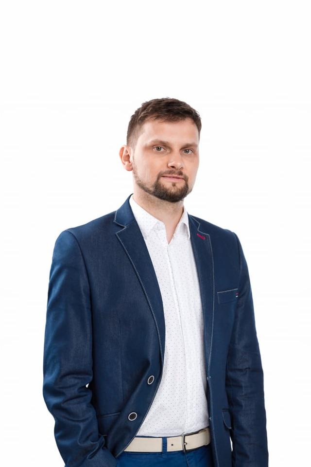 Portret mężczyzny z krótką brodą, ubranego w białą koszulę w kropki i niebieską marynarkę z białymi przeszyciami, pozującego na białym tle.