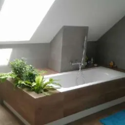 Jasna łazienka na poddaszu ze skośnym sufitem, biała wanna obudowana drewnianą konstrukcją z wbudowaną donicą z zielonymi roślinami, szare płytki na ścianach, błękitny ręcznik i dywanik.