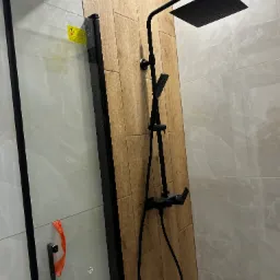 Nowoczesny zestaw natryskowy w czarnym kolorze zamontowany na ścianie wyłożonej płytkami imitującymi drewno, widoczna kabina prysznicowa z czarnymi profilami, fragment naklejki ostrzegawczej...