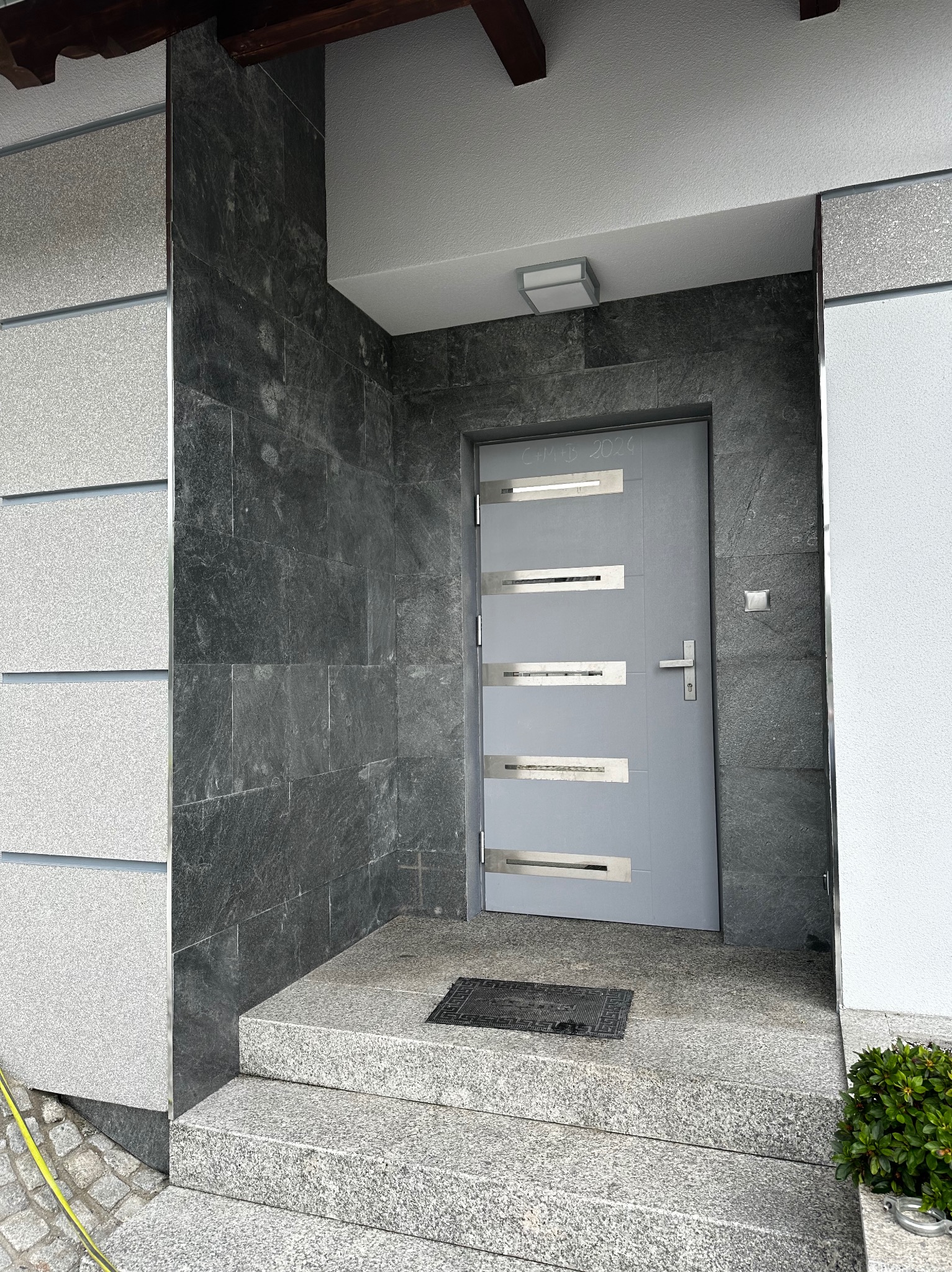 Szare drzwi wejściowe z poziomymi, metalowymi akcentami, otoczone ciemnoszarą okładziną z kamienia naturalnego, jasnoszarym tynkiem oraz schodami z jasnego granitu. Nad drzwiami widoczny napis...