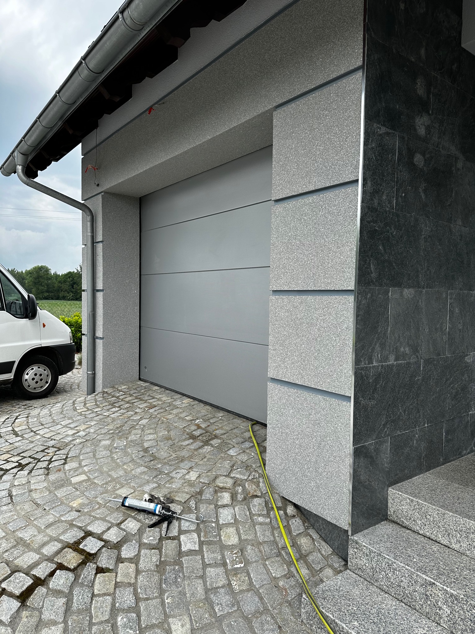 Szara brama garażowa segmentowa, obok wykończona elewacja z szarego kamienia, na brukowanym podjeździe leży pistolet do silikonu i żółty kabel.