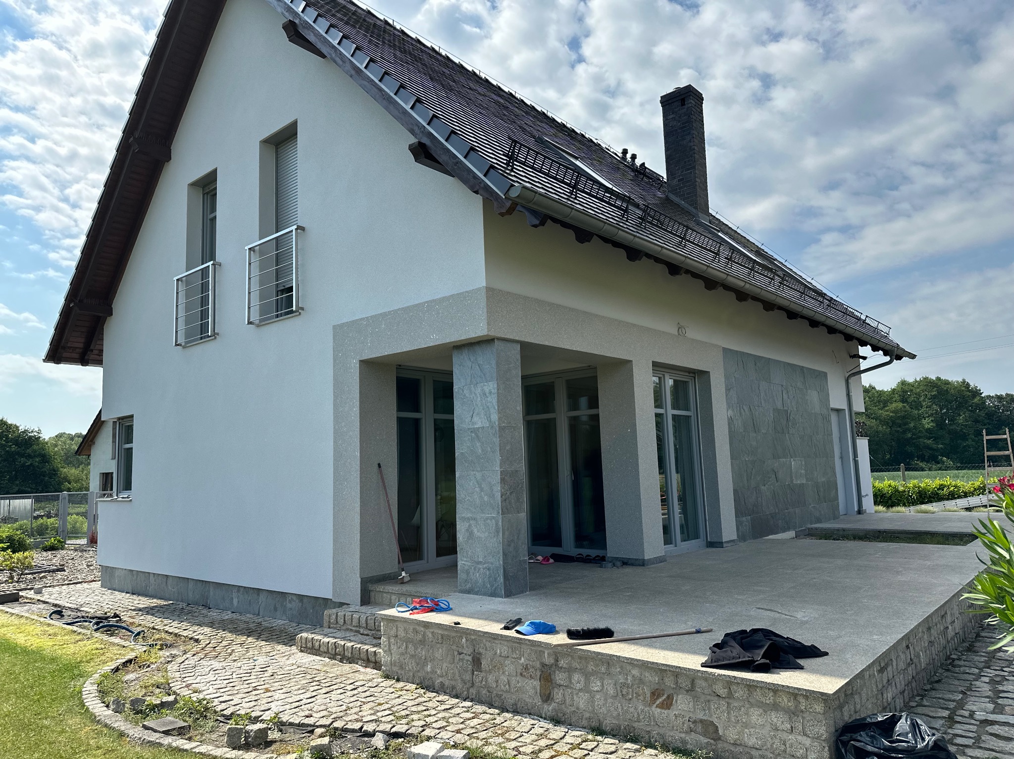 Biały dom z szarą elewacją i kamienną podmurówką, widoczne narzędzia i akcesoria malarskie na tarasie, częściowo wykończony teren wokół domu