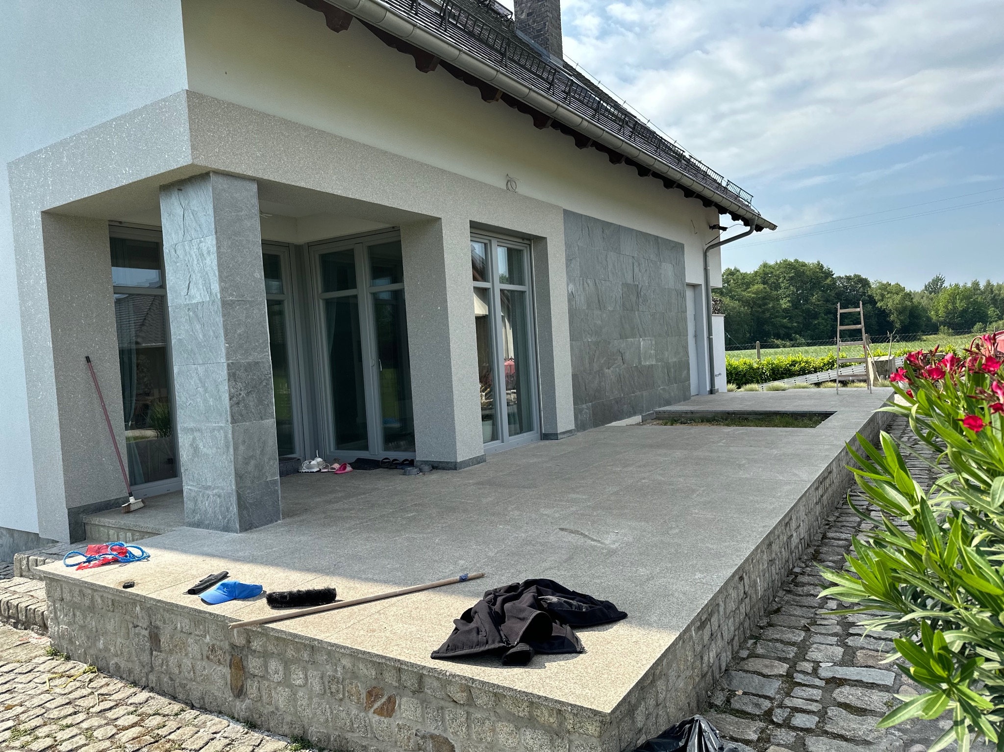 Wykończona elewacja domu z szarymi płytami kamiennymi, widoczne narzędzia i akcesoria po zakończonych pracach, fragment tarasu i zieleni.