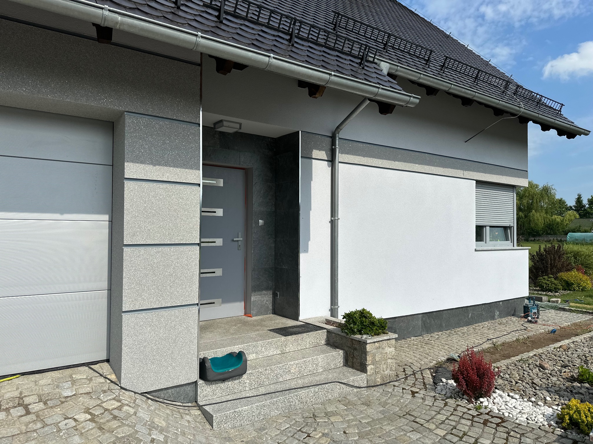 Nowoczesny dom jednorodzinny z elewacją w odcieniach szarości, z garażem i wejściem, widoczne schody wejściowe z donicą i chodnik z kostki brukowej.