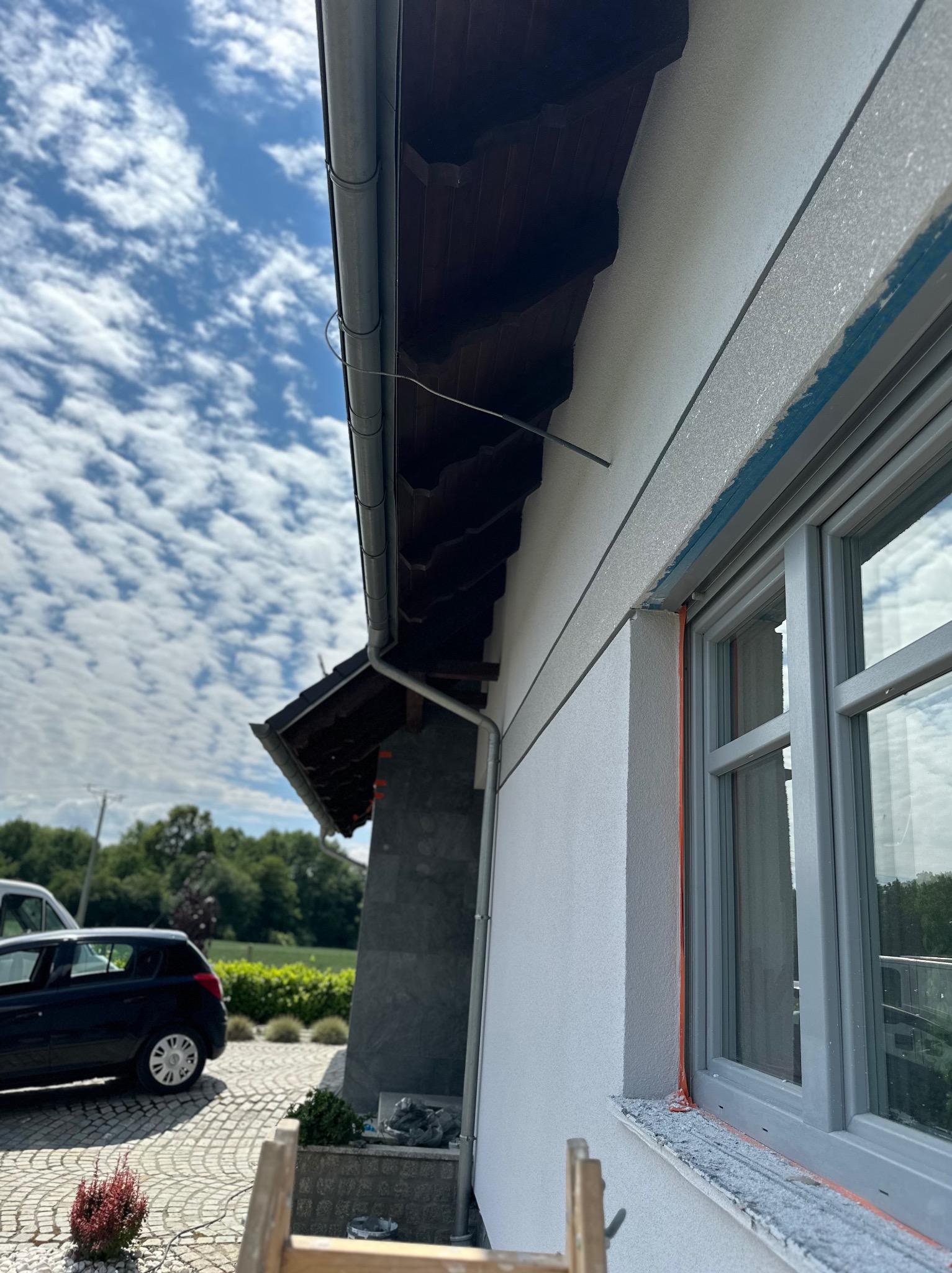 Fragment elewacji domu z oknem w szarej ramie, widoczne prace wykończeniowe, zabezpieczone folią i z resztkami zaprawy na parapecie, w tle błękitne niebo z chmurami i fragment dachu z rynną.