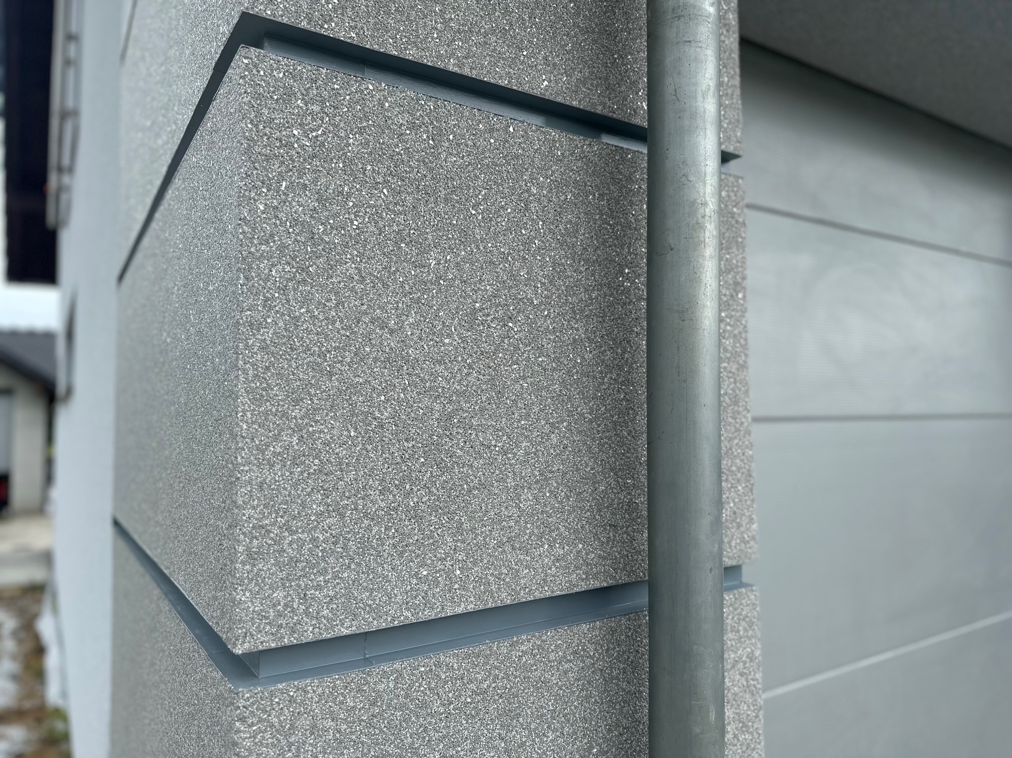 Szary narożnik elewacji budynku z fakturą baranka, z widocznym metalowym profilem i rurą spustową.