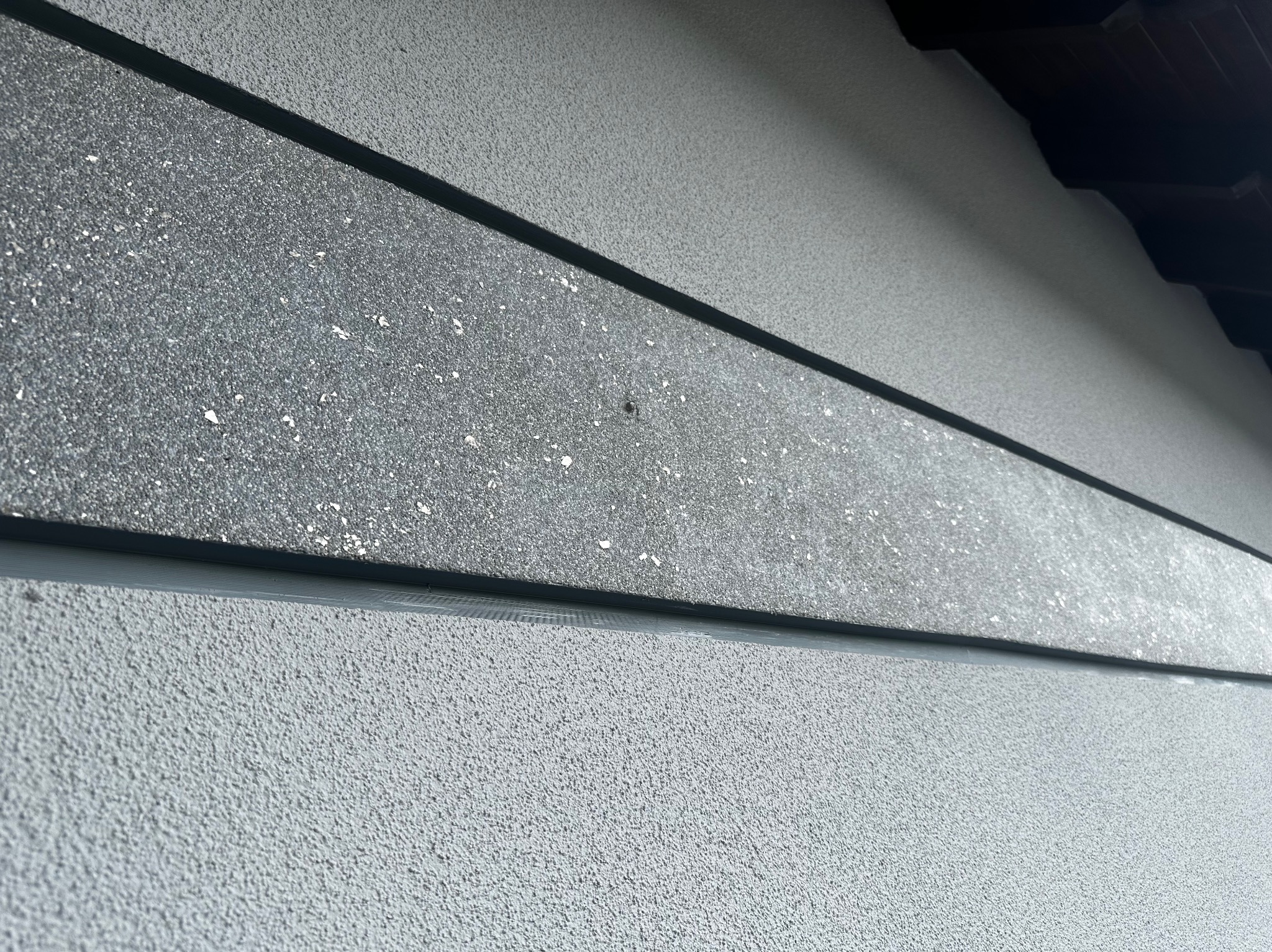 Fragment elewacji budynku z widoczną strukturą tynku i elementami dekoracyjnymi w odcieniach szarości, z fragmentem dachu w górnym rogu.