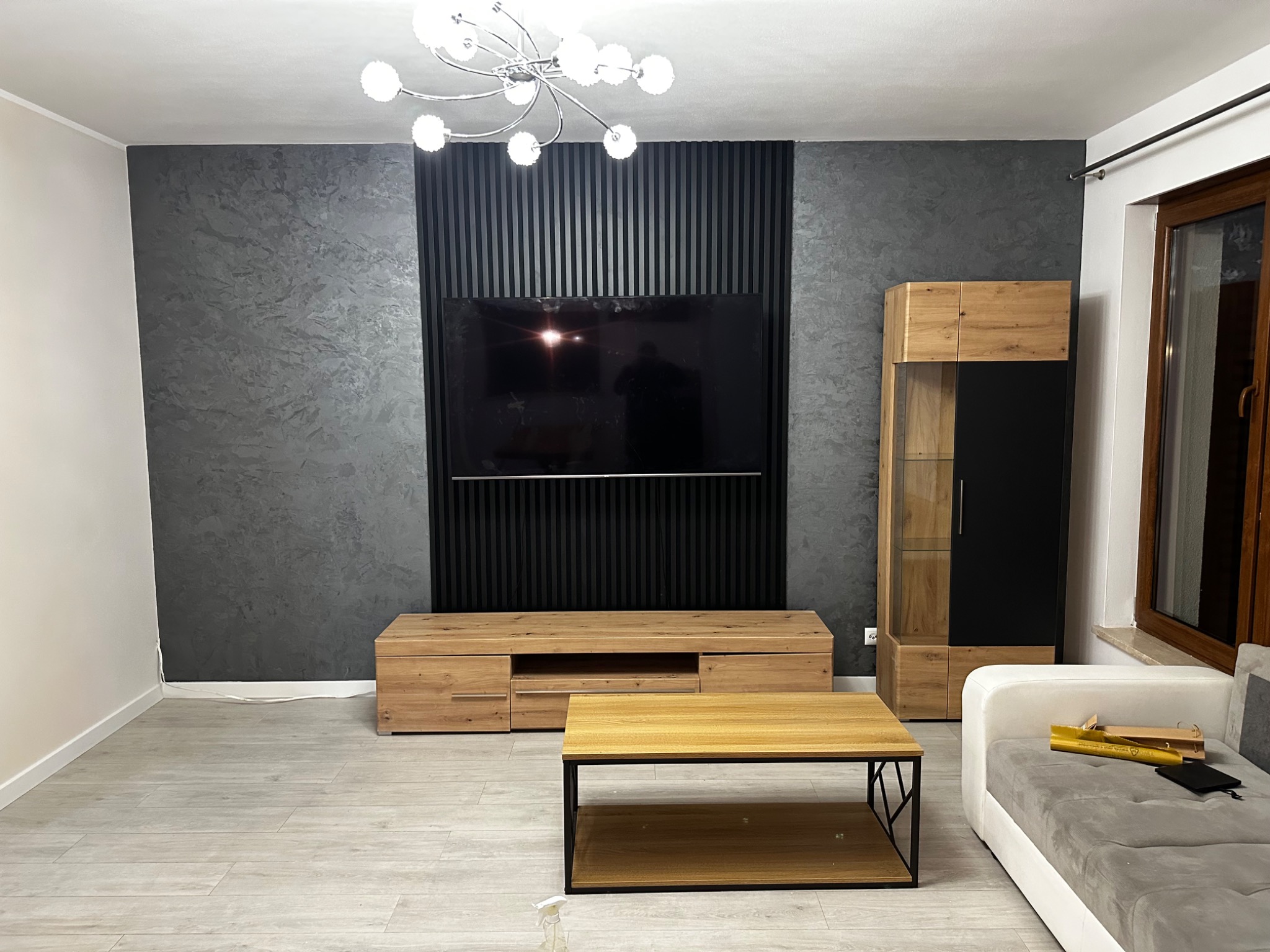 Nowoczesny salon z szarą ścianą o dekoracyjnej fakturze, czarnymi pionowymi panelami za telewizorem, drewnianą szafką RTV i stolikiem kawowym, oraz białym narożnikiem z żółtymi elementami...