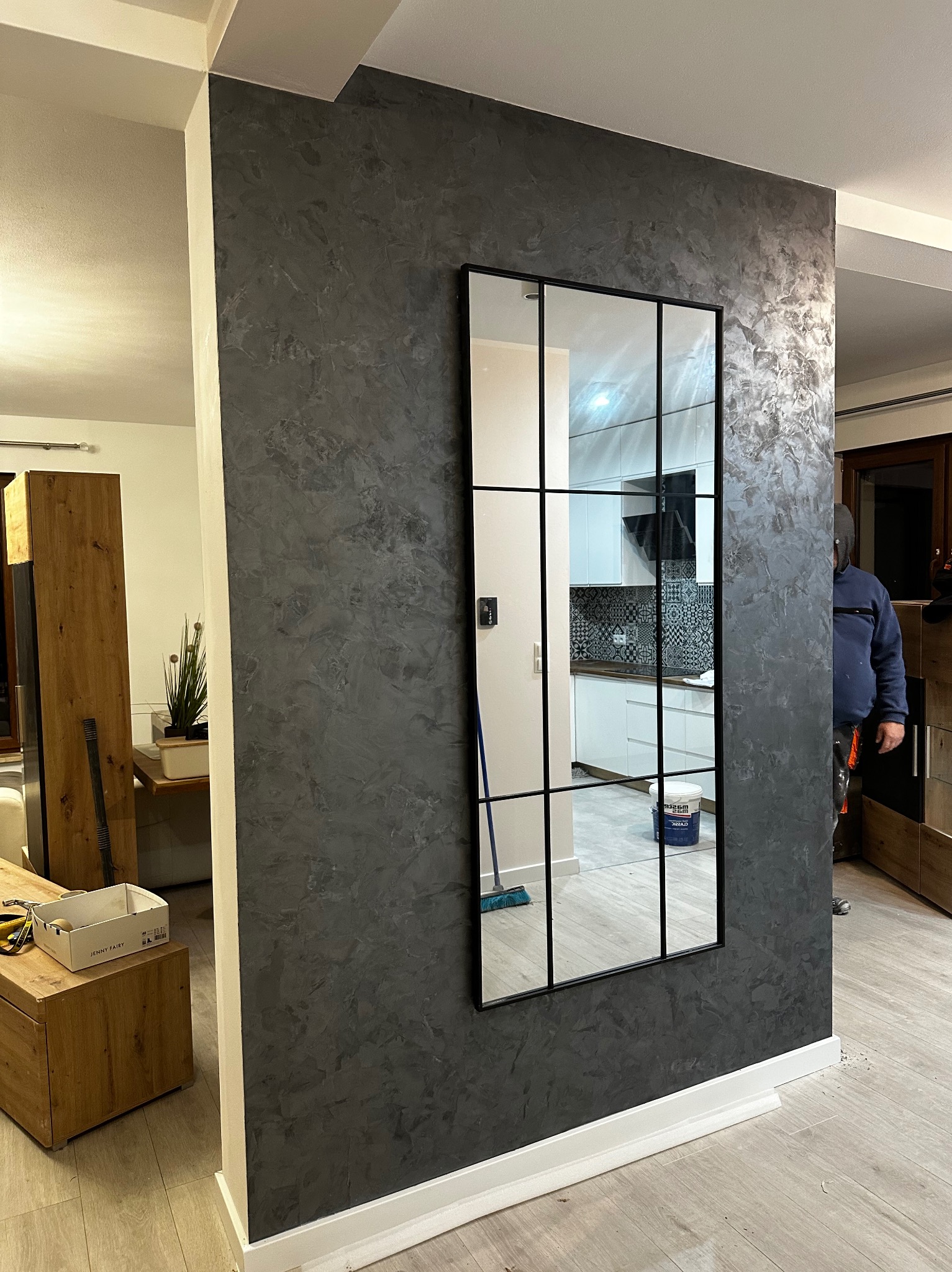 Ściana wykończona dekoracyjnym tynkiem w odcieniach szarości, z zamontowanym lustrem w czarnej ramie, odbijającym wnętrze kuchni z białą zabudową i czarno-białą mozaiką. W tle widoczna osoba...