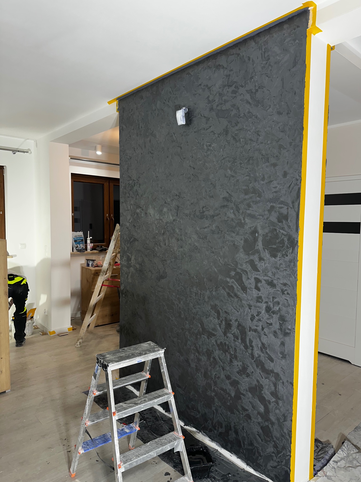 Ściana z dekoracyjną, ciemną strukturą, zabezpieczona żółtą taśmą malarską, drabina malarska w pomieszczeniu w trakcie prac wykończeniowych.