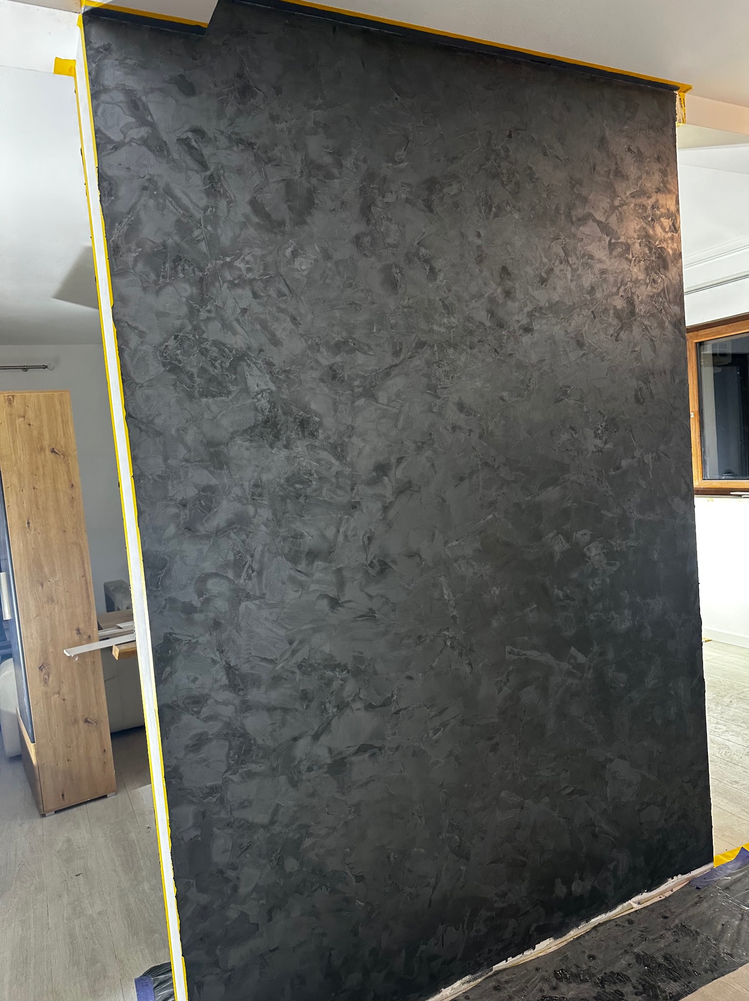 Czarna ściana z dekoracyjną strukturą, zabezpieczona żółtą taśmą malarską przy suficie i podłodze, widoczne elementy wyposażenia wnętrza w tle.