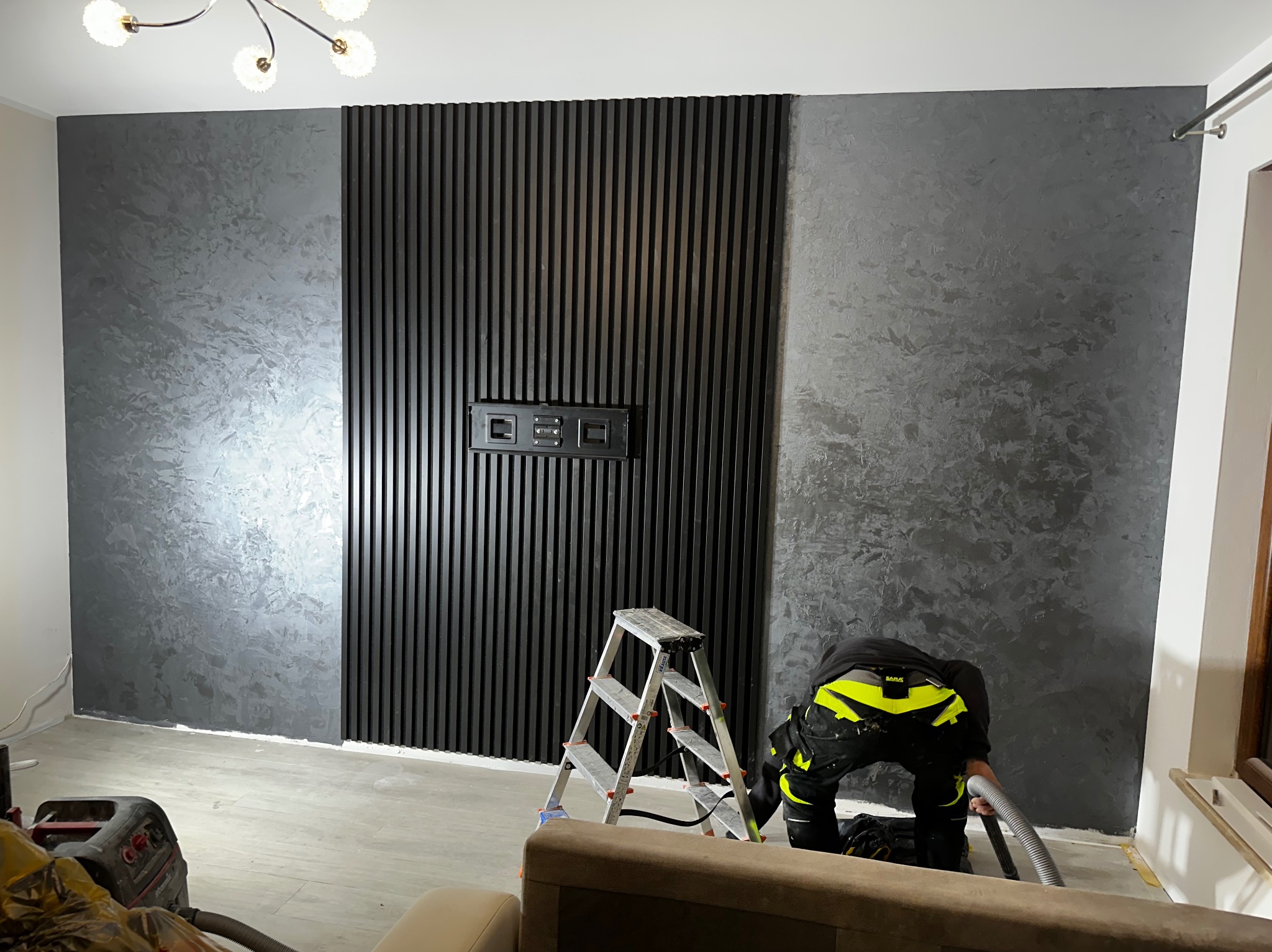 Szara ściana z dekoracyjną strukturą i panelem z czarnych lameli; pracownik na drabinie odkurza podłogę.