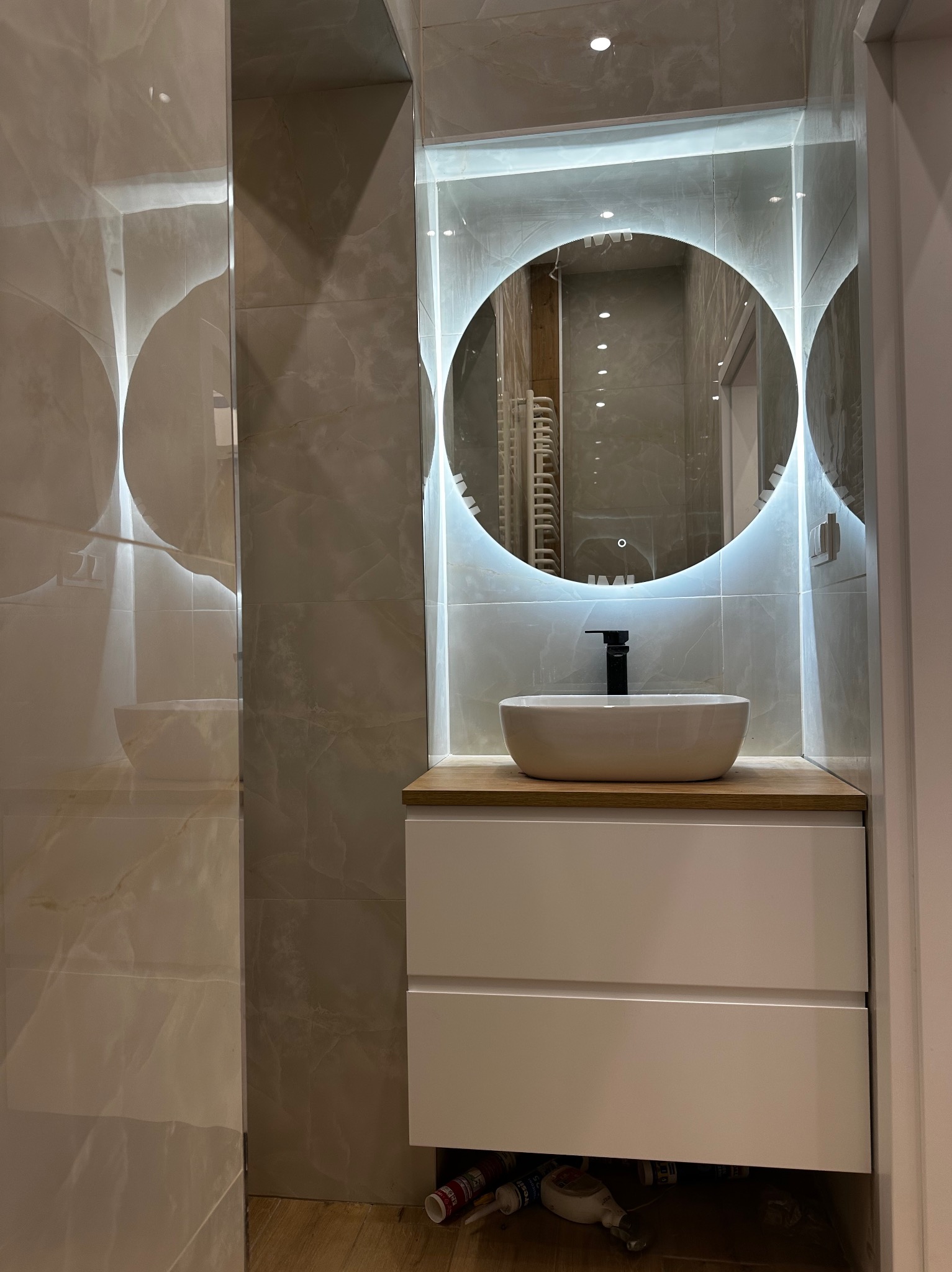 Nowoczesna łazienka z okrągłym lustrem podświetlanym LED, umywalką nablatową i szafką z szufladami, wykończona płytkami imitującymi marmur.